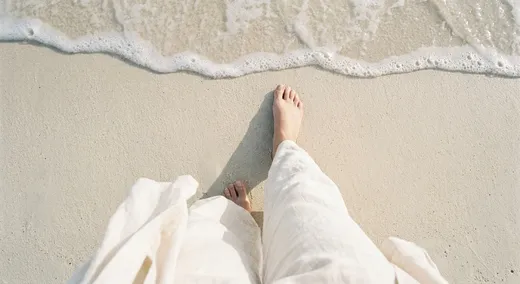 Nackte Füße in weißer Leinenhose stehen im auslaufenden Wellenschaum am Sandstrand