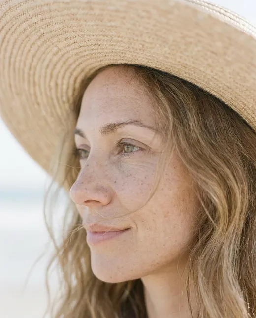 Porträt einer Frau mit Sonnenhut im Profil, Blick zur Seite, verschwommener Strand im Hintergrund