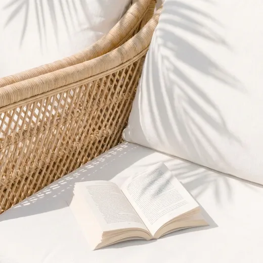 Aufgeschlagenes Buch auf weißen Kissen neben einem Rattankorb, Schatten von Palmblättern im Sonnenlicht